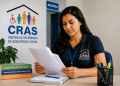 Mulher de cabelo castanho lendo papéis no CRAS com livros e crachá azul sobre a mesa, simbolizando o funcionamento dos concursos com vagas para o CRAS.