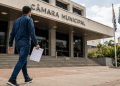 Candidato caminha em direção à Câmara Municipal segurando um documento de concurso público.
