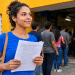 Jovem candidata segura ficha de inscrição em frente a escola com fila de pessoas para concurso público.