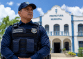 Agente uniformizado da Guarda Municipal em frente ao prédio de uma prefeitura, com palmeiras ao redor