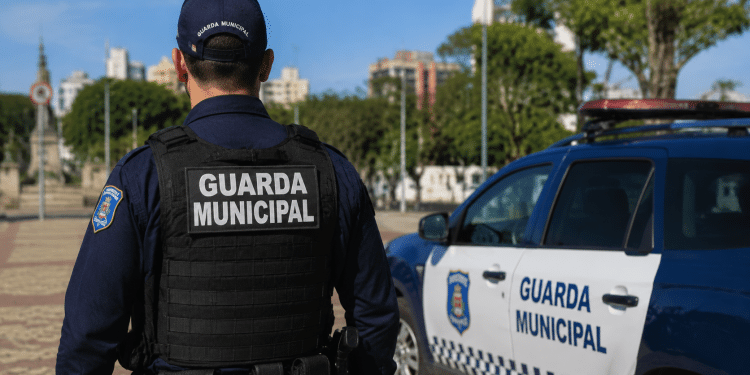 Guarda Municipal de costas em patrulhamento ao lado de viatura oficial em área urbana.
