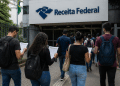 Pessoas com mochilas e documentos de concurso público caminhando em direção ao prédio da Receita Federal no Brasil.