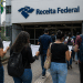 Pessoas com mochilas e documentos de concurso público caminhando em direção ao prédio da Receita Federal no Brasil.