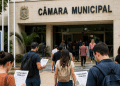 Pessoas se dirigindo a uma Câmara Municipal com documentos de concurso público em mãos.