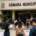 Pessoas se dirigindo a uma Câmara Municipal com documentos de concurso público em mãos.