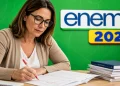 Professora corrigindo redação do Enem 2026 em mesa de trabalho, com materiais de avaliação e slogan ao fundo.