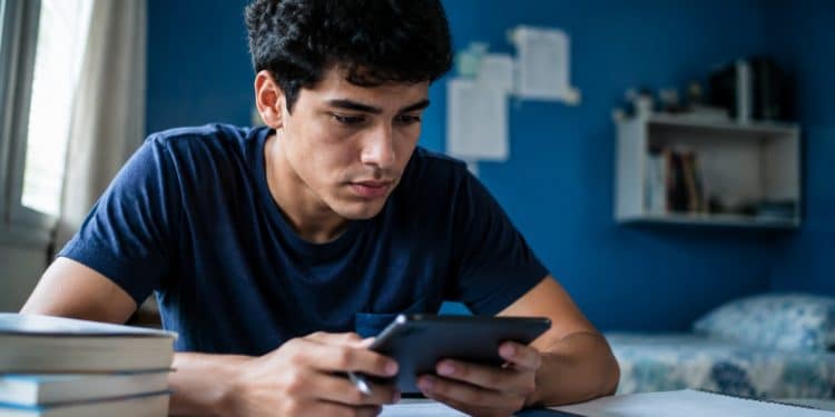 Estudante consulta o celular à mesa, com livros e caderno, para verificar informações do Encceja 2026 na Página do Participante.