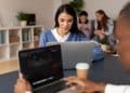 Jovem de suéter azul estuda em notebook em ambiente compartilhado com outros estudantes ao fundo