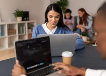 Jovem de suéter azul estuda em notebook em ambiente compartilhado com outros estudantes ao fundo