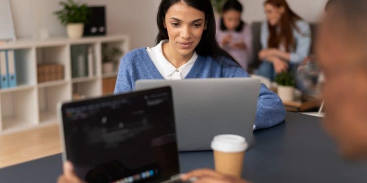 Jovem de suéter azul estuda em notebook em ambiente compartilhado com outros estudantes ao fundo