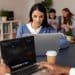 Jovem de suéter azul estuda em notebook em ambiente compartilhado com outros estudantes ao fundo