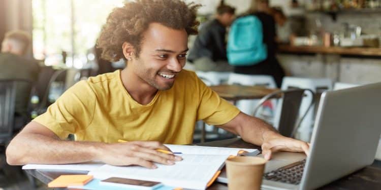 Estudante sorridente estuda em notebook com caderno e anotações, situação que remete aos cursos livres usados como horas complementares na faculdade