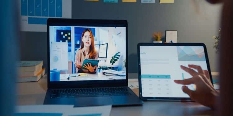 Jovem ministra aula online em notebook enquanto pessoa analisa gráficos em tablet no escritório, simbolizando os cursos livres para o currículo.