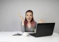 Mulher jovem sorridente com cabelo rosa segura lápis ao lado de notebook em mesa branca, simbolizando as opções de curso para ganhar dinheiro online.