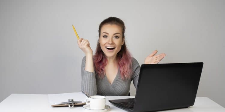Mulher jovem sorridente com cabelo rosa segura lápis ao lado de notebook em mesa branca, simbolizando as opções de curso para ganhar dinheiro online.