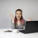 Mulher jovem sorridente com cabelo rosa segura lápis ao lado de notebook em mesa branca, simbolizando as opções de curso para ganhar dinheiro online.