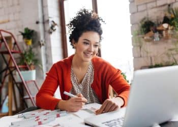 Mulher de casaco vermelho sorrindo enquanto faz anotações diante de um notebook