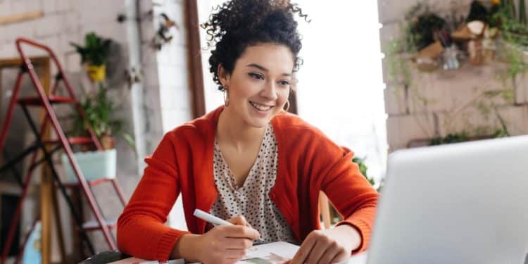 Mulher de casaco vermelho sorrindo enquanto faz anotações diante de um notebook
