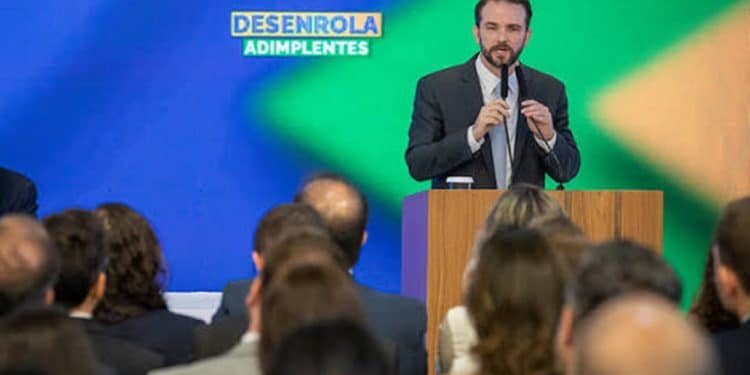 Homem de terno discursa em púlpito diante de plateia sob painel do Desenrola Adimplentes em evento do governo