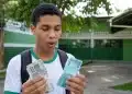 Estudante segura notas de dinheiro em frente a uma escola, com expressão de surpresa, representando a possibilidade de pagamento duplicado do Pé-de-Meia em um mês.