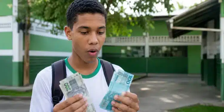 Estudante segura notas de dinheiro em frente a uma escola, com expressão de surpresa, representando a possibilidade de pagamento duplicado do Pé-de-Meia em um mês.