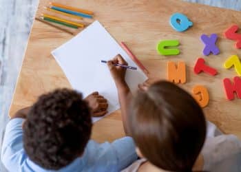 Criança escreve em folha de papel ao lado de professora, com letras coloridas e lápis sobre a mesa