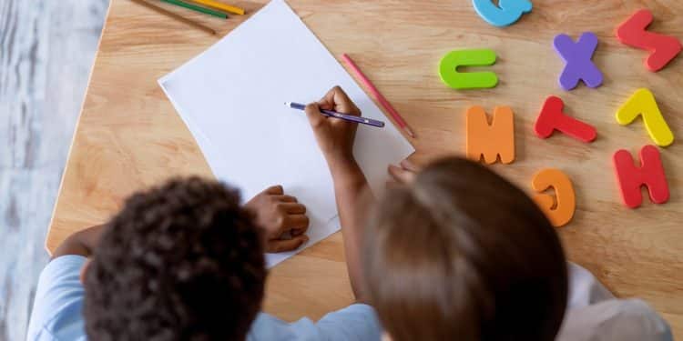 Criança escreve em folha de papel ao lado de professora, com letras coloridas e lápis sobre a mesa
