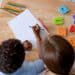 Criança escreve em folha de papel ao lado de professora, com letras coloridas e lápis sobre a mesa