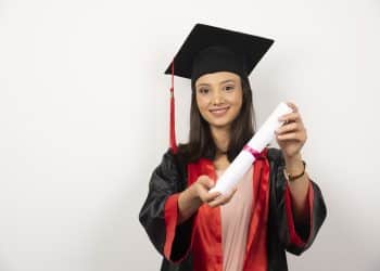 Jovem estudante sorrindo com beca preta e detalhes vermelhos segura diploma enrolado em destaque, simbolizando o prazo final para pedir atendimento especializado no Enade 2026.
