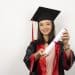 Jovem estudante sorrindo com beca preta e detalhes vermelhos segura diploma enrolado em destaque, simbolizando o prazo final para pedir atendimento especializado no Enade 2026.