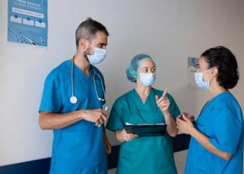 Três profissionais de saúde de máscara conversando em um corredor, um deles segurando prancheta