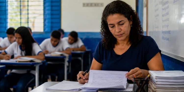 Professora corrige atividades em sala de aula da rede pública, ilustrando os salários dos docentes da rede estadual e o estado que mais paga.