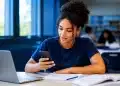 Jovem universitária consulta informações sobre a inscrição no Fies pelo celular, com notebook e documentos sobre a mesa.