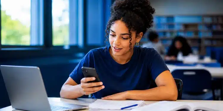 Jovem universitária consulta informações sobre a inscrição no Fies pelo celular, com notebook e documentos sobre a mesa.
