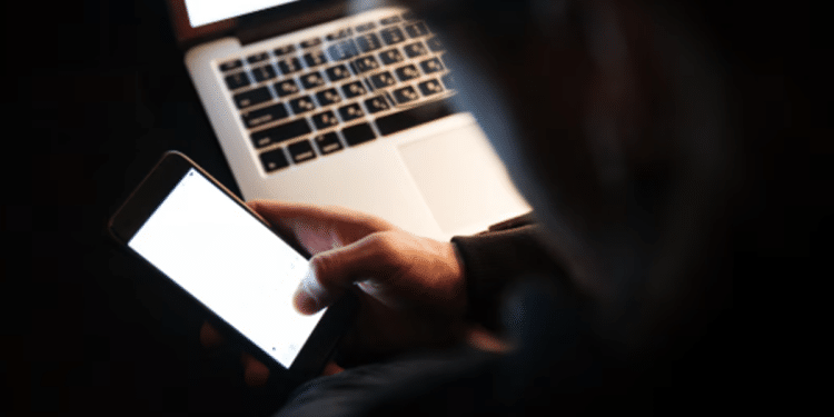 Pessoa segurando um celular iluminado diante de um notebook em ambiente escuro