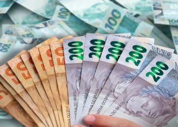 Mão segurando um leque de notas de real de 50, 100 e 200 reais, com mais dinheiro ao fundo, cena que representa o piso salarial pago às carreiras alcançadas pela lei.