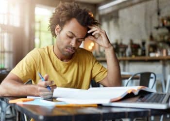 Jovem de camiseta amarela estudando de forma concentrada com livro, caderno e notebook