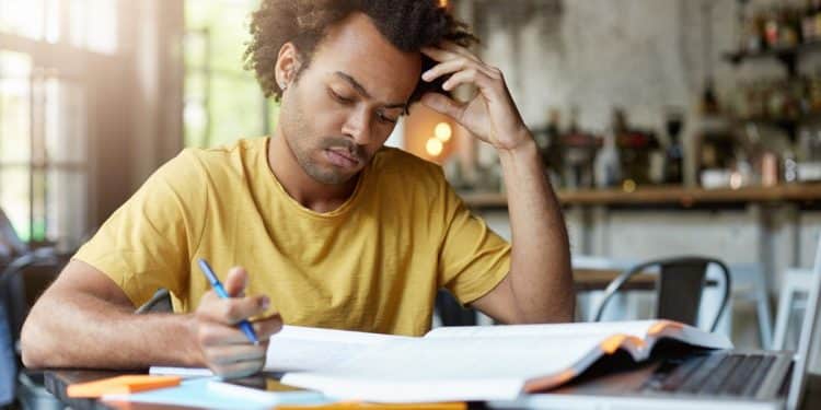 Jovem de camiseta amarela estudando de forma concentrada com livro, caderno e notebook
