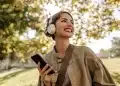 Mulher sorridente ouve música com fones de ouvido enquanto segura o celular em um parque.