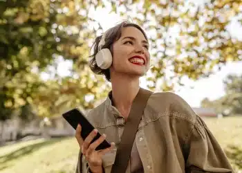 Mulher sorridente ouve música com fones de ouvido enquanto segura o celular em um parque.