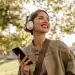 Mulher sorridente ouve música com fones de ouvido enquanto segura o celular em um parque.