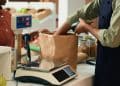 Comerciante pesando um produto em uma balança no caixa de um mercado, com cliente ao lado