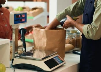 Comerciante pesando um produto em uma balança no caixa de um mercado, com cliente ao lado