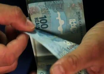 Pessoa conta cédulas de 100 reais, representando o pagamento do INSS liberado nesta sexta-feira, 3 de julho.