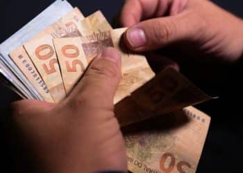 Pessoa contando várias cédulas de cinquenta reais sobre fundo escuro