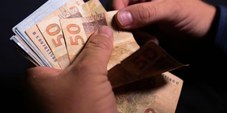 Pessoa contando várias cédulas de cinquenta reais sobre fundo escuro