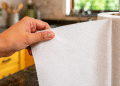 Mão destaca uma folha de papel-toalha do rolo em um suporte, com a cozinha ao fundo