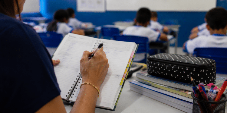 Professora fazendo anotações em agenda durante aula em sala de ensino fundamental, ilustrando investimentos do Fundeb na educação.
