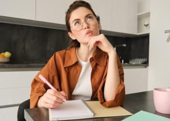 Mulher de óculos com expressão pensativa, segurando caneta diante de um caderno aberto