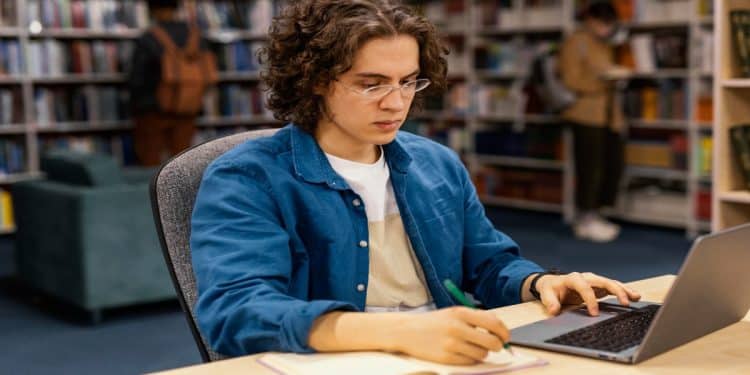 Imagem ilustrativa de candidato estudando com notebook e caderno em uma biblioteca, representando a preparação para o concurso INSS 2026 e as 8.501 vagas pedidas para o cargo de nível médio.